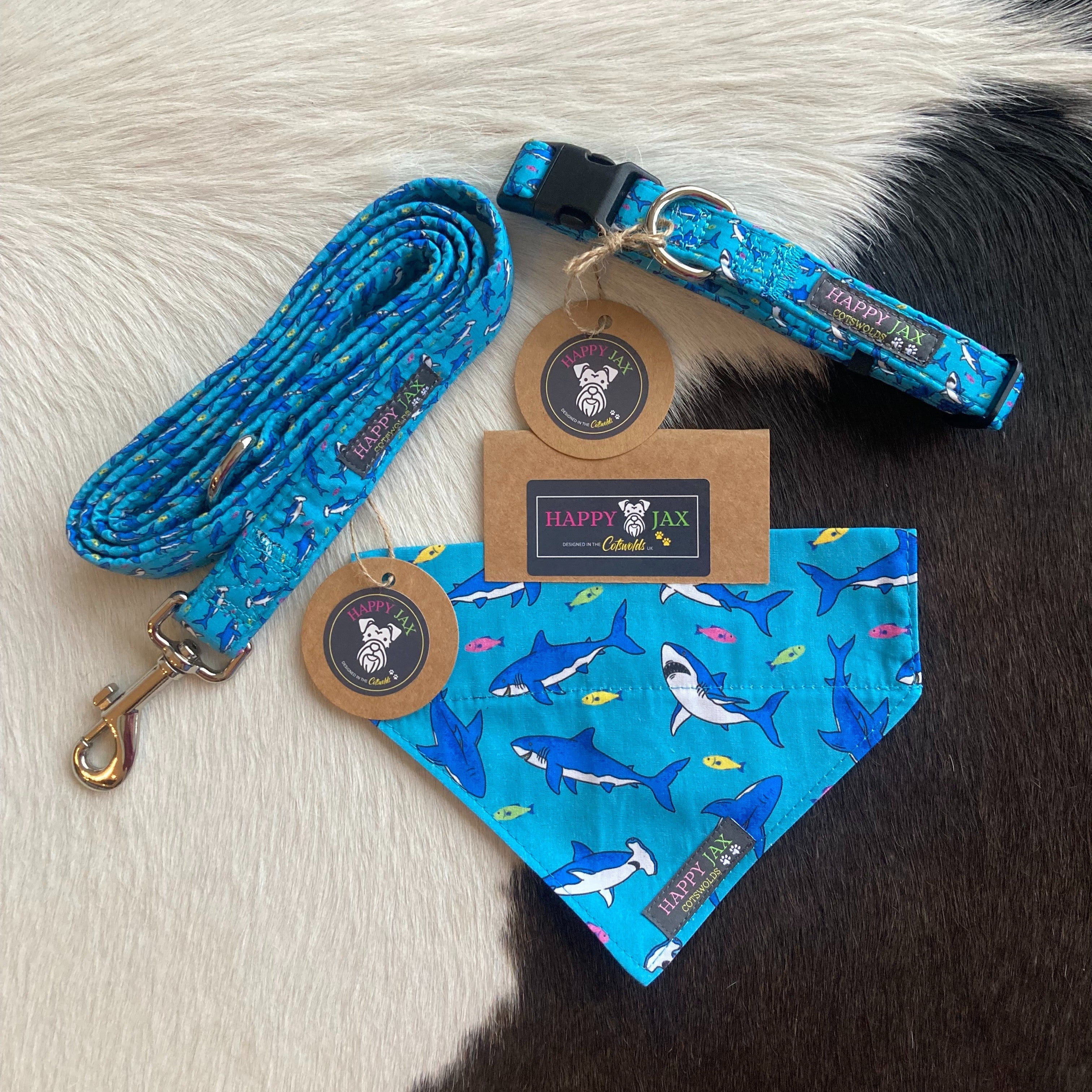 Bundle with an animal collar, bandana and lead in a bright and colourful design of dark blue regular and hammer head sharks surrounded by multicoloured fish on a bright blue background. Products are suitable for both cats and dogs and available in a range of sizes, made with cotton and designed in the Cotswolds.